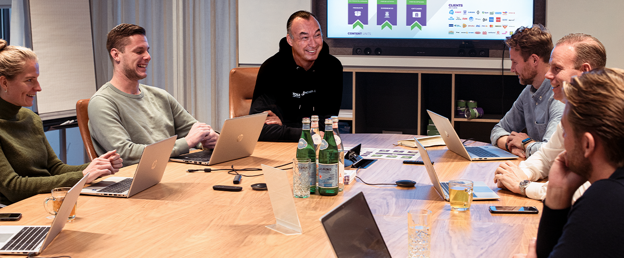 Teamoverleg met lachende collega’s rond een vergadertafel met laptops terwijl zij samenwerken aan strategie, ideeën en klantoplossingen.
